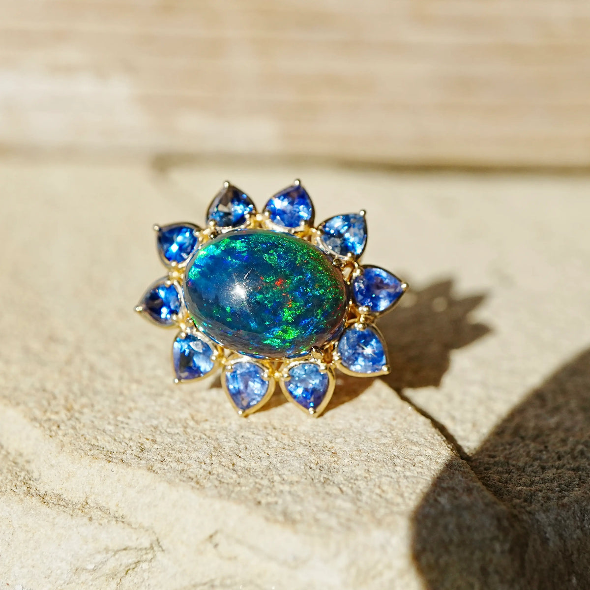 A 14k gold ring featuring a large oval iridescent black opal center surrounded by a halo of blue sapphires, photographed on a textured beige stone surface with warm sunlight and shadows.