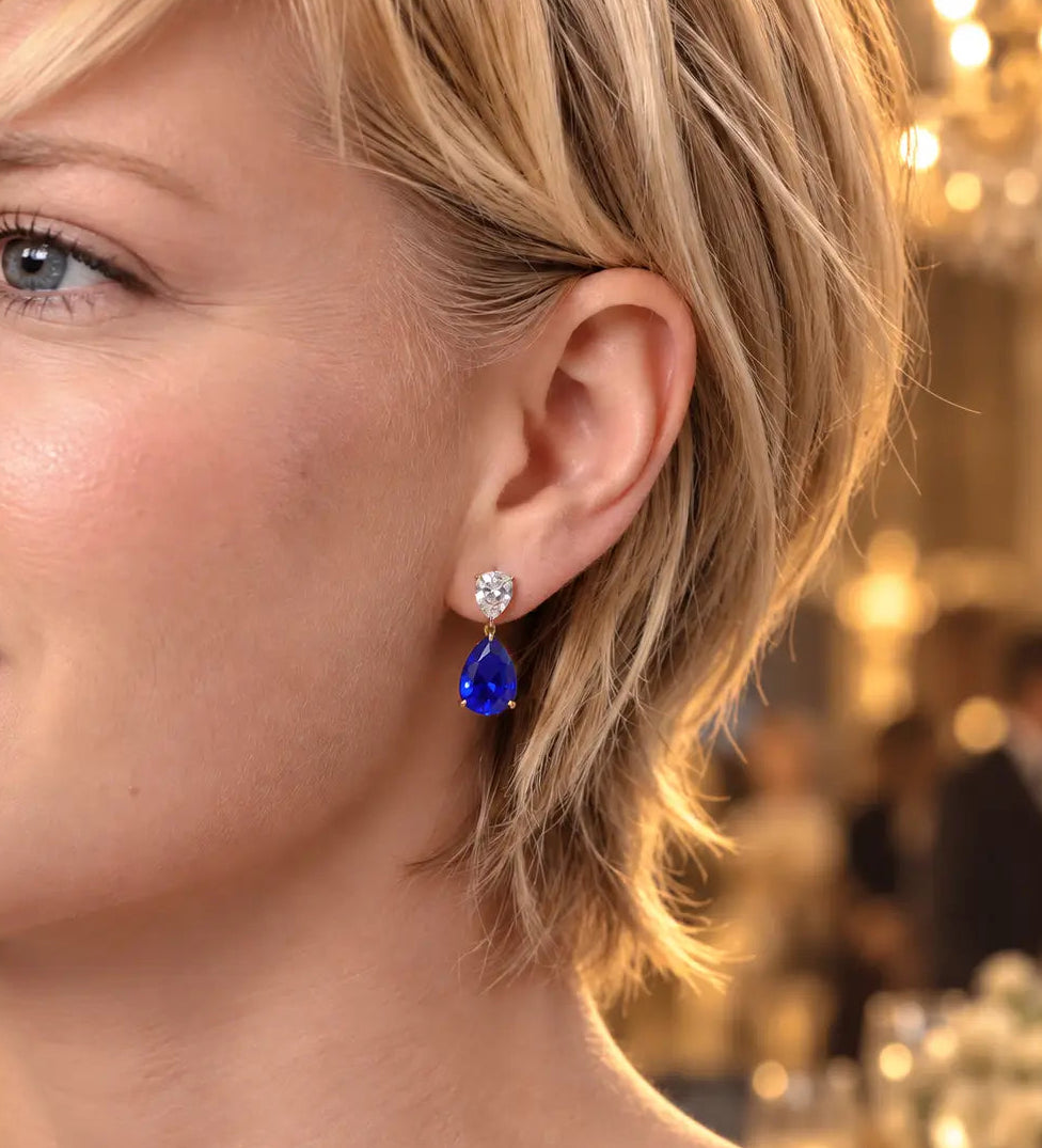 A close-up of a woman with short blonde hair wearing a teardrop-shaped blue gemstone dangle earring with a small clear stone above it, set against a softly blurred indoor background with warm lights.